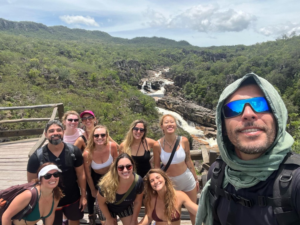 Grupo de turistas sorrindo com o guia Diego Navi em mirante do Parque Nacional da Chapada dos Veadeiros, com cachoeira e cerrado ao fundo