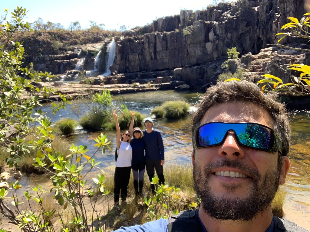 Guide and visitors taking a selfie by the river with a waterfall in the background, Chapada dos Veadeiros